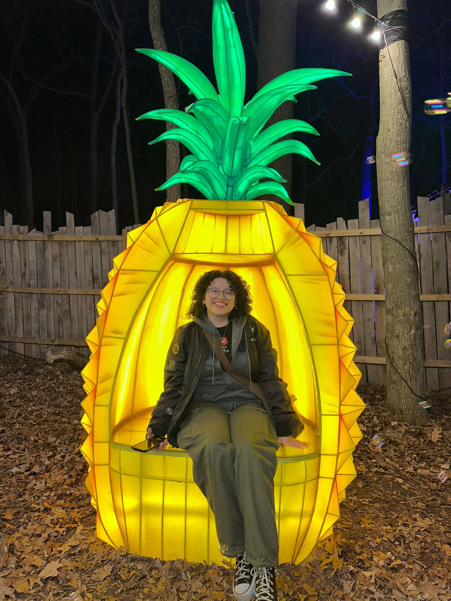 Atticus with black, curly, shoulder-length hair and glasses, sits on a glowing yellow pineapple
